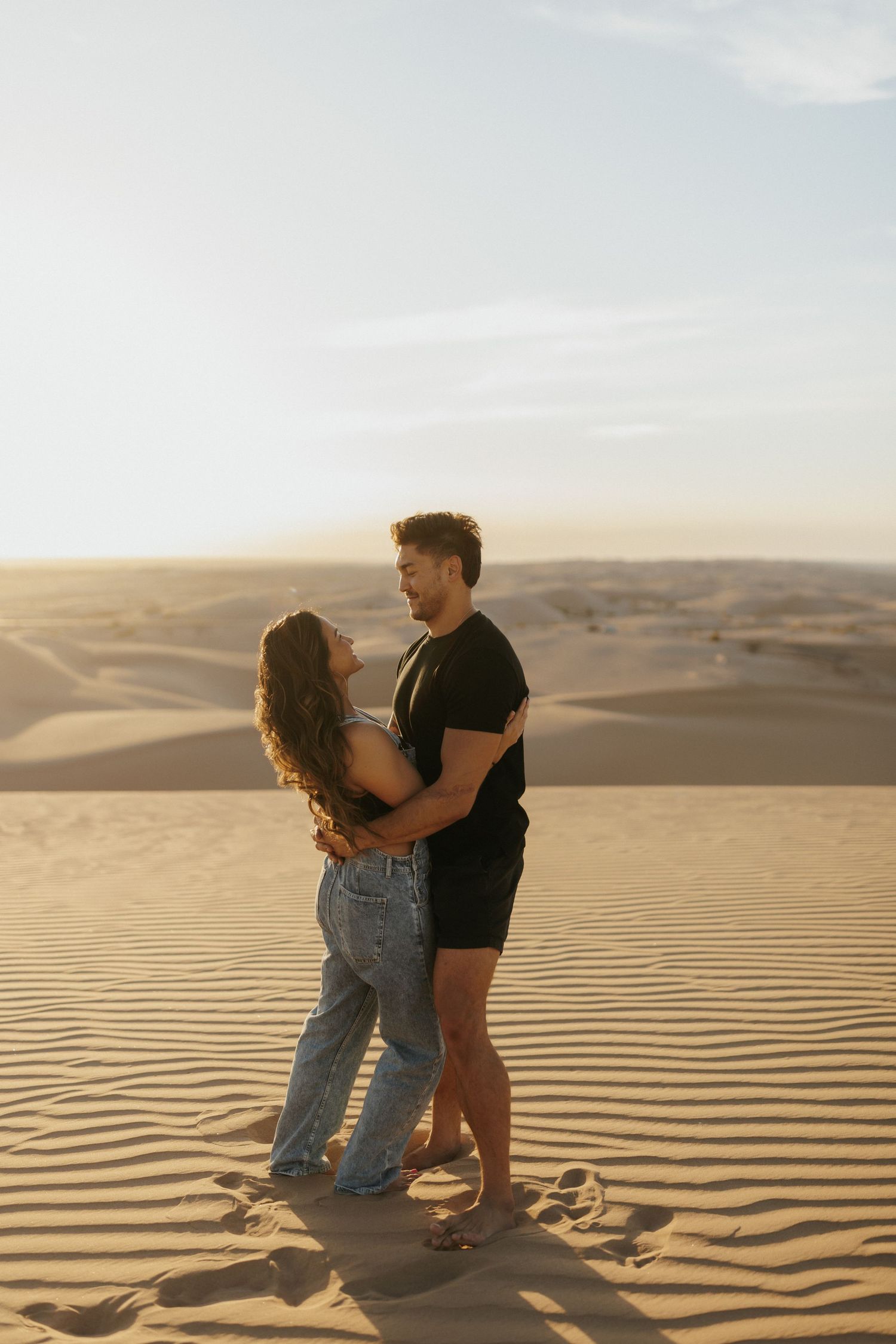 Two people sit together on rippling sand dunes at sunset in a desert landscape.