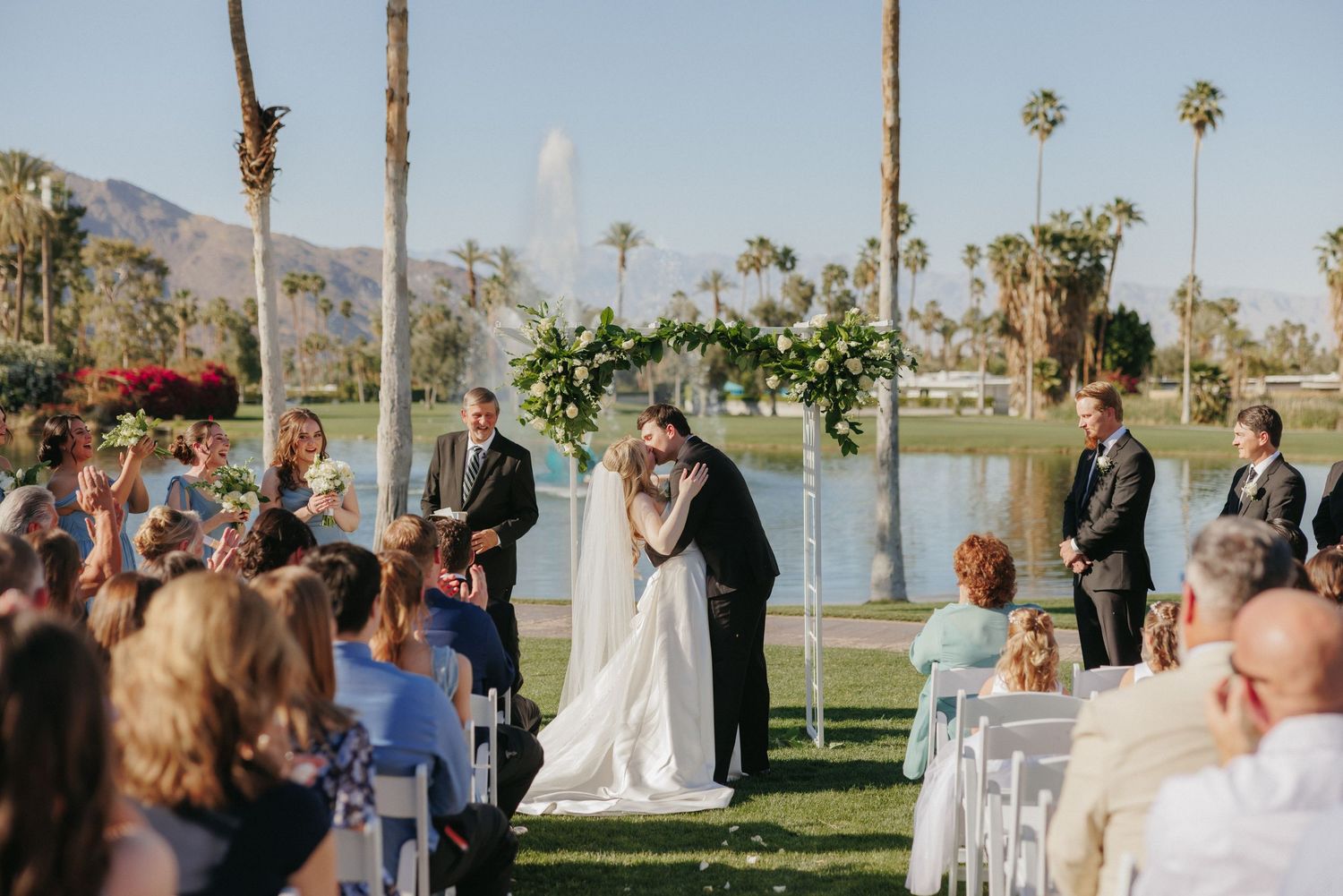 Bride and groom share first kiss at outdoor wedding ceremony by a pond with floral arch.