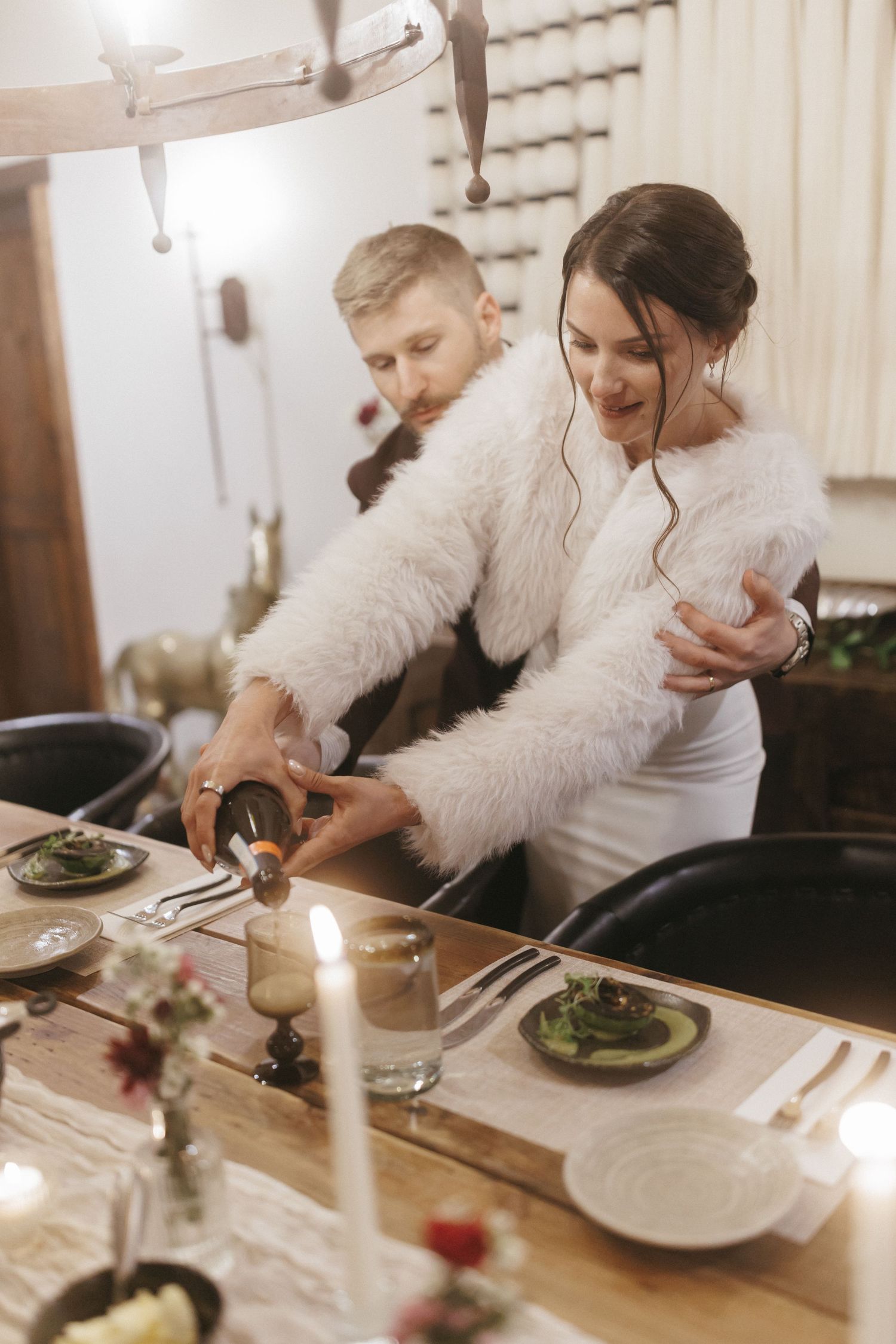 A smiling bride in a white fur jacket cuts cake with groom at a candlelit reception.
