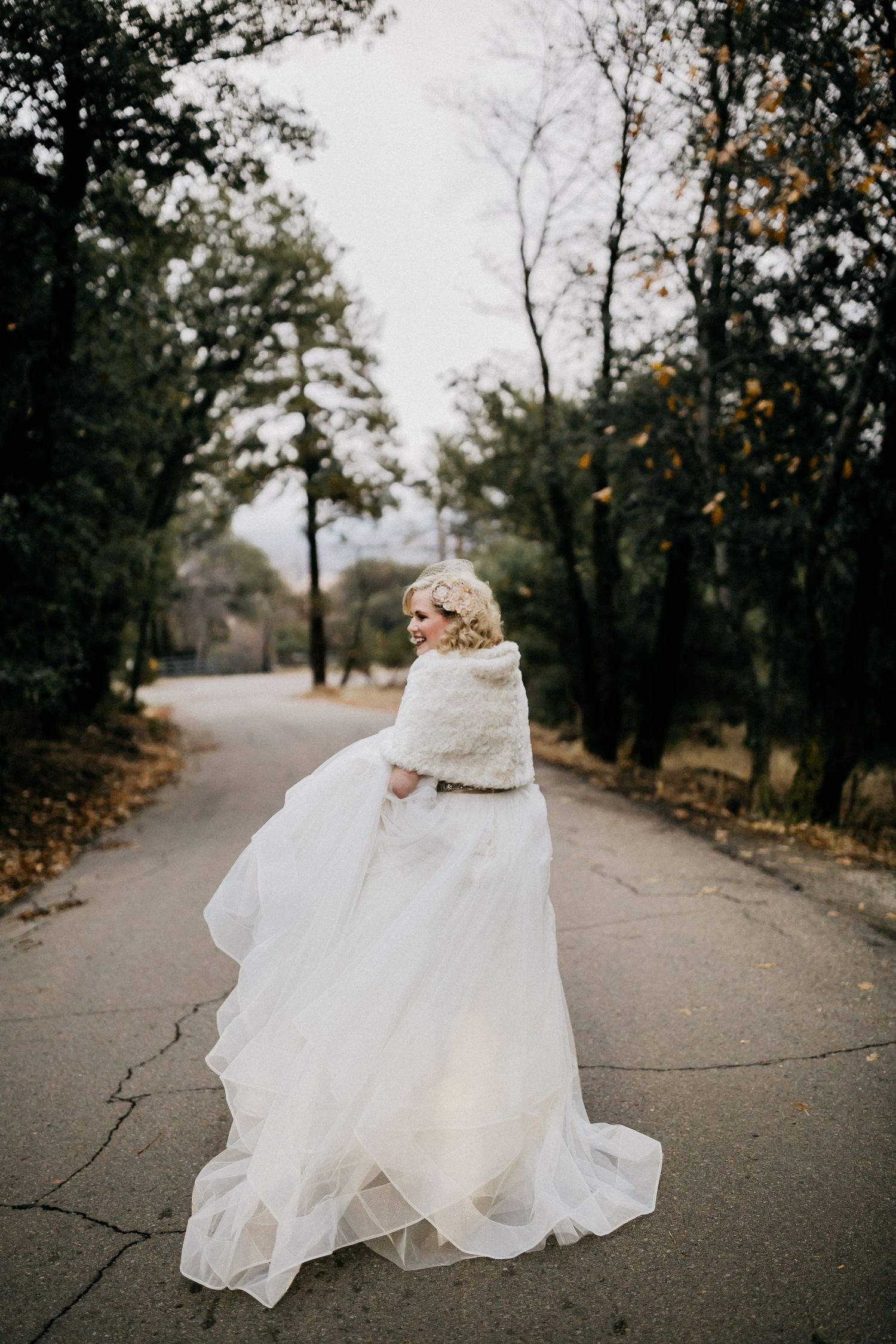 A flowing white wedding dress twirls dramatically on a paved path surrounded by autumn trees.