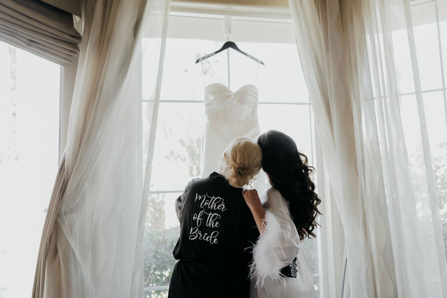 A wedding dress hangs in front of a bright window with sheer white curtains creating a romantic silhouette.