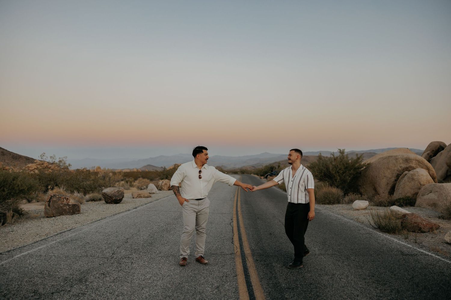Two people standing on an empty desert road at dusk surrounded by large boulders in a mountainous landscape.
