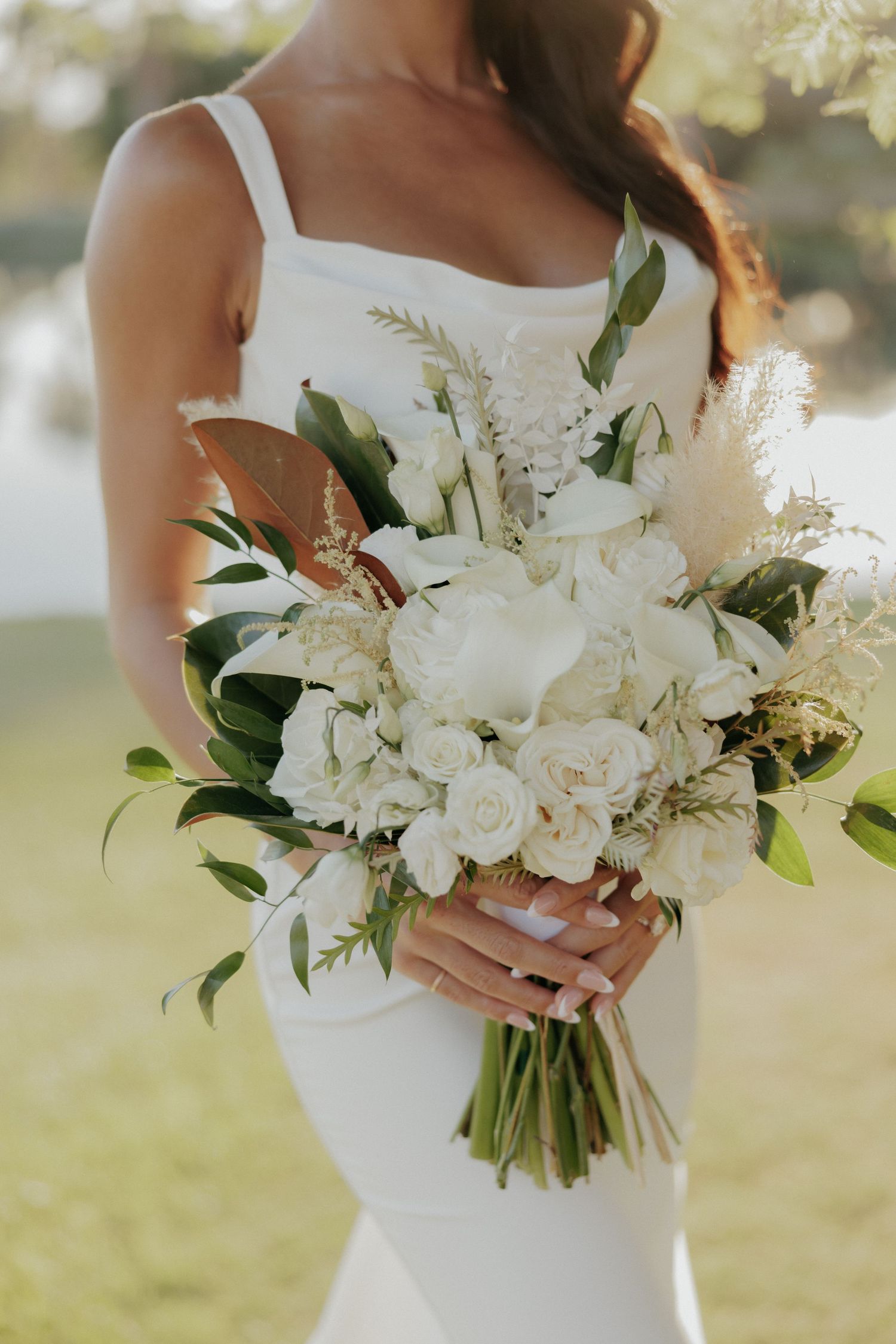 Elegant white bridal bouquet featuring peonies and greenery held against a white wedding dress in natural sunlight.