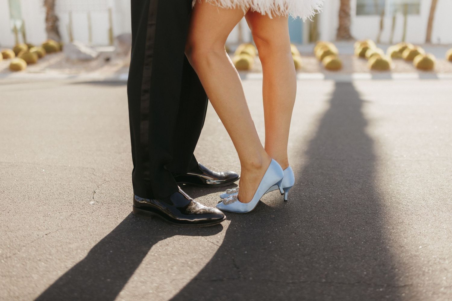 Close-up of bride and groom's feet, she wearing blue heels and he in black dress shoes, standing on pavement.