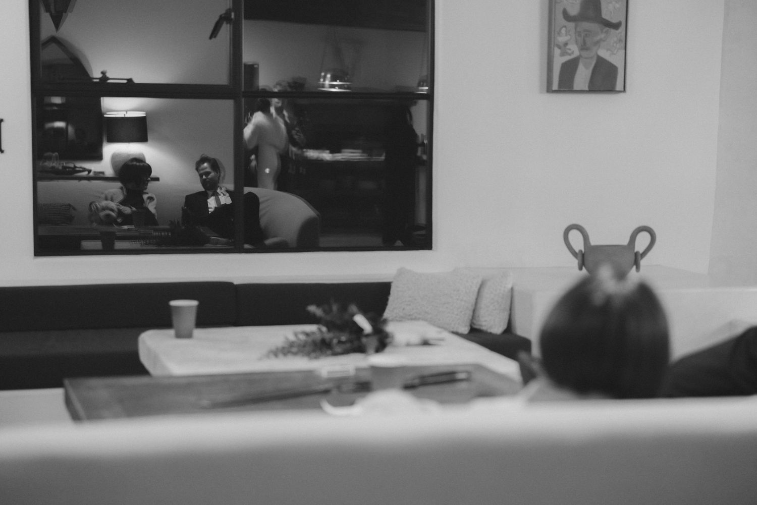 Black and white photo of a living room with people reflected in a window behind sofa.