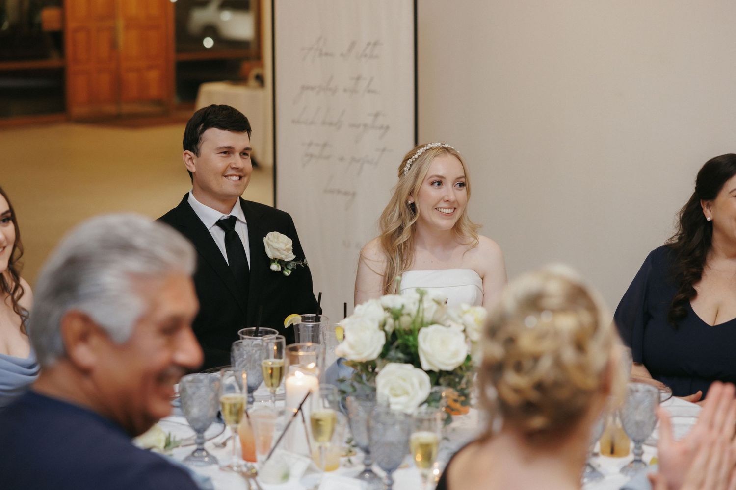 Smiling bride and groom sit at sweetheart table during elegant wedding reception.