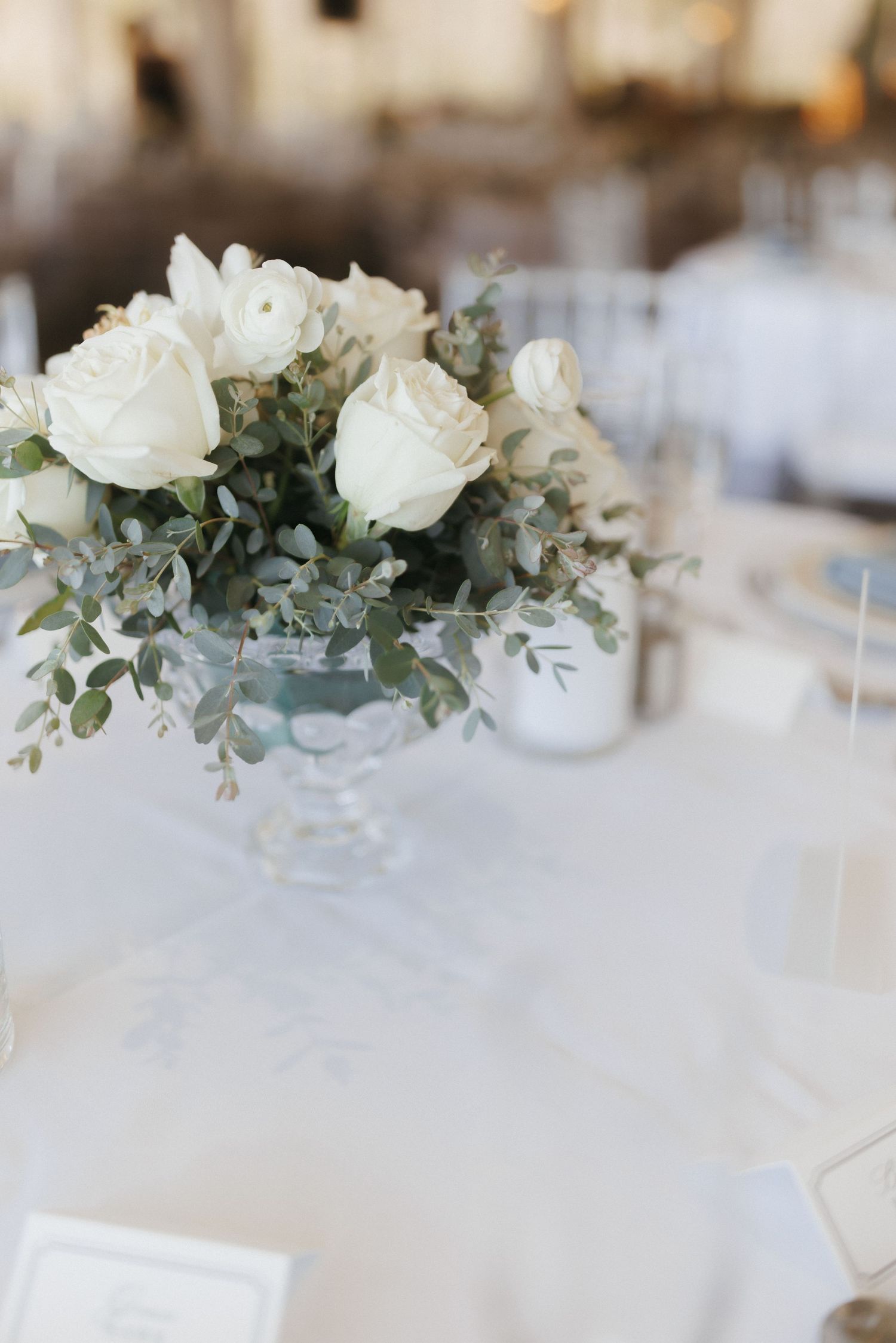 Low floral centerpiece of white roses and eucalyptus in crystal compote vase on white tablecloth.