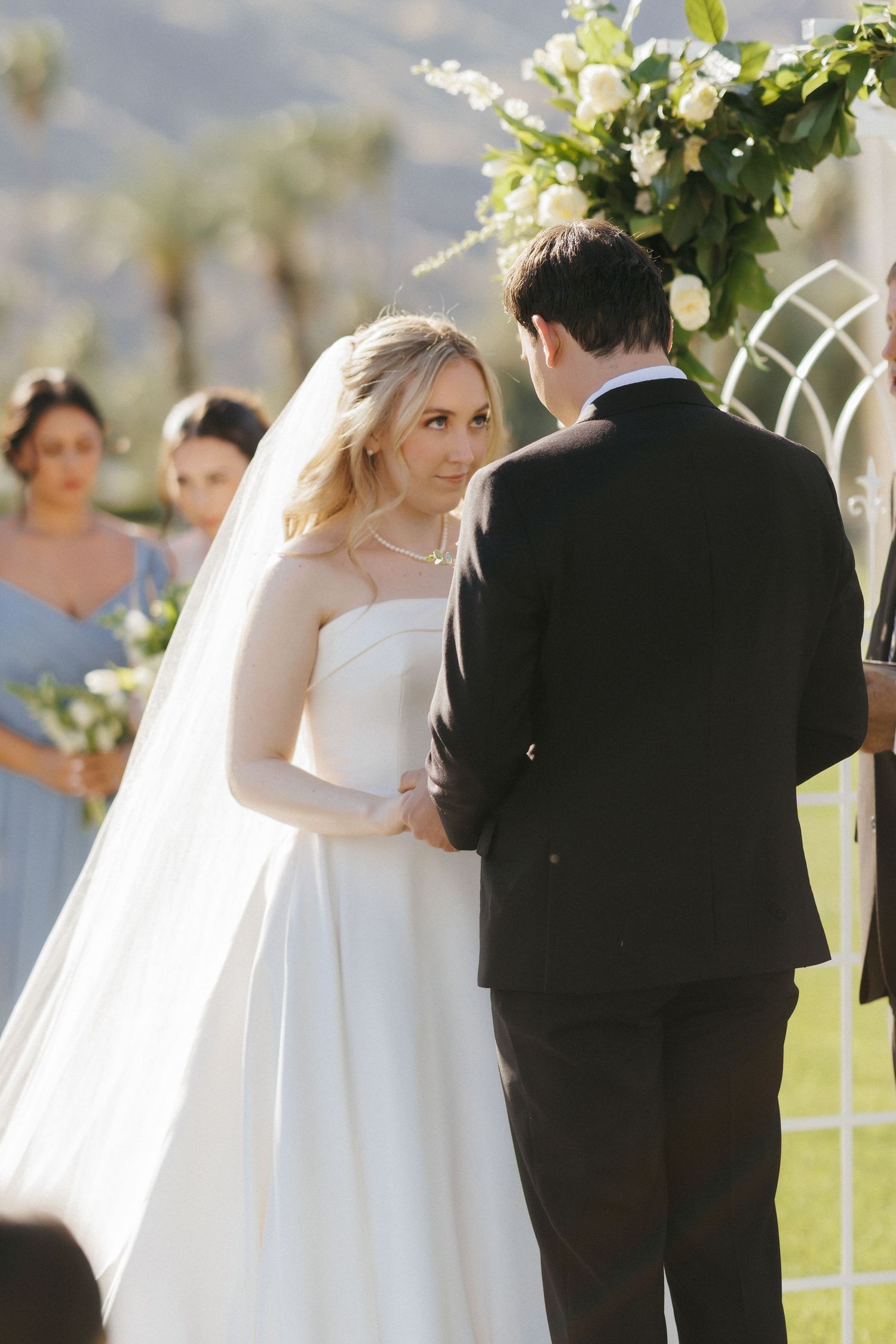 Bride in a white gown exchanging vows with groom under a floral arch at an outdoor wedding.