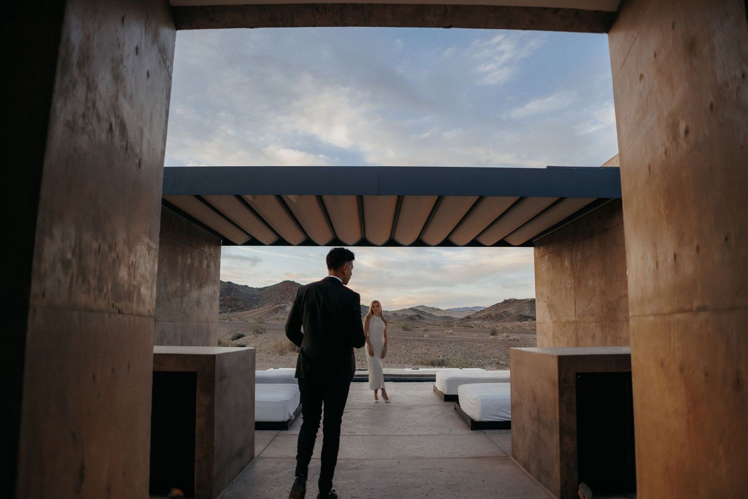 Couple in formal attire walks through modern desert architecture toward scenic mountain views at dusk.