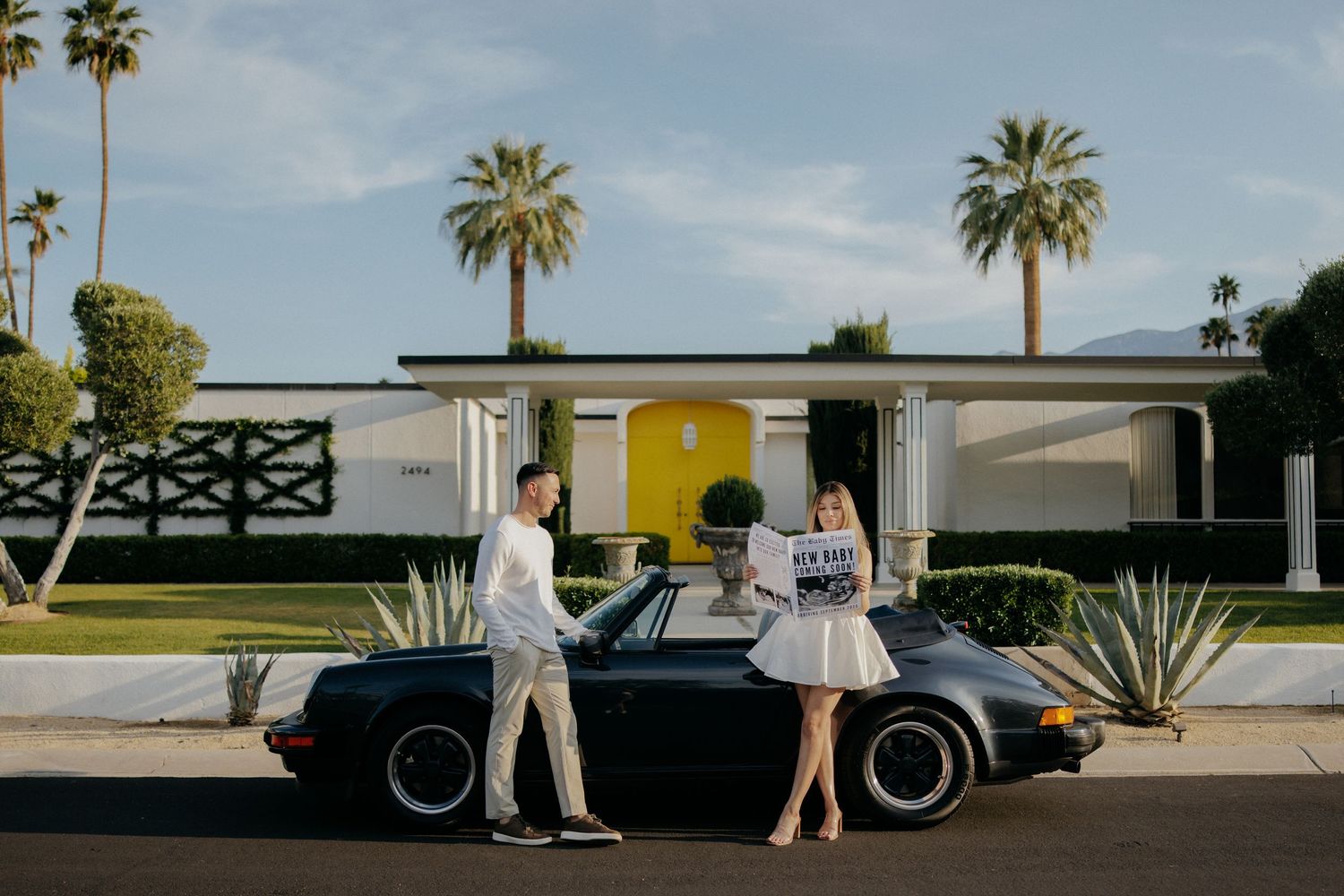 Couple posing with a black Porsche convertible in front of a mid-century modern home in Palm Springs.