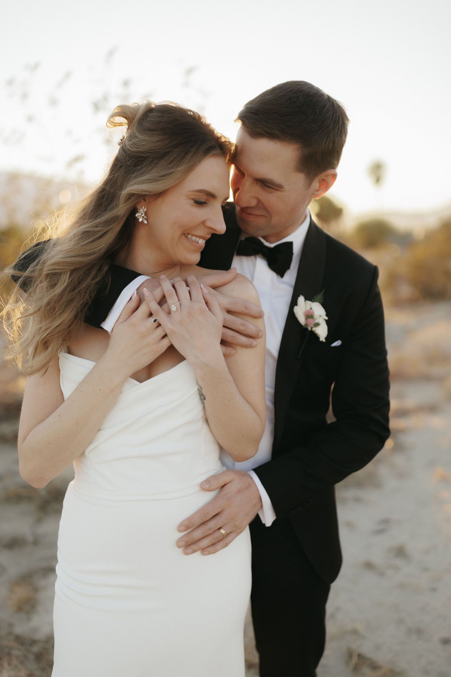 Bride and groom embracing outdoors in desert setting, she wears white gown, he wears black tuxedo with boutonniere.