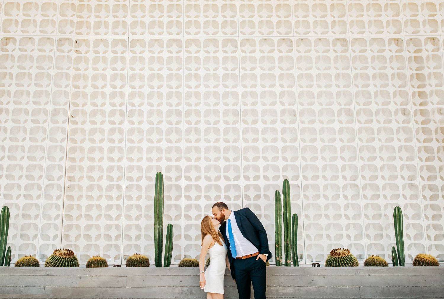 A couple shares a romantic moment surrounded by tall desert cactus plants in an outdoor garden setting.