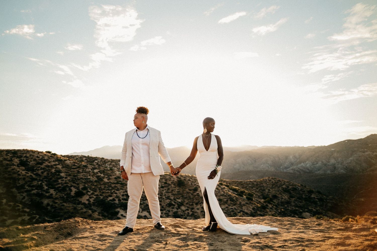 Newlyweds in white outfits walking along a scenic mountain ridge at dusk.