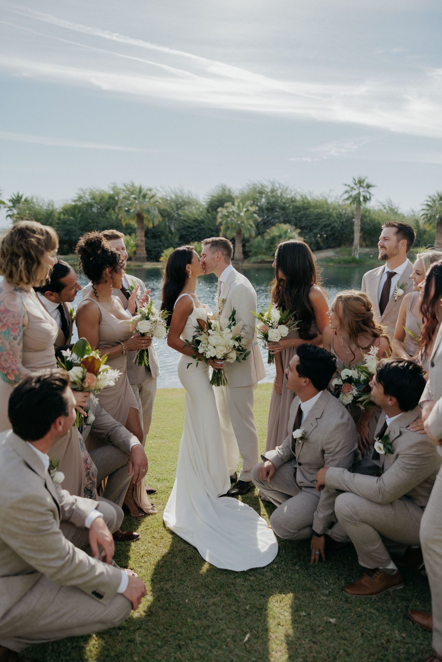 Wedding party celebrates with bridesmaids in blush dresses and groomsmen in tan suits during an outdoor lawn reception.