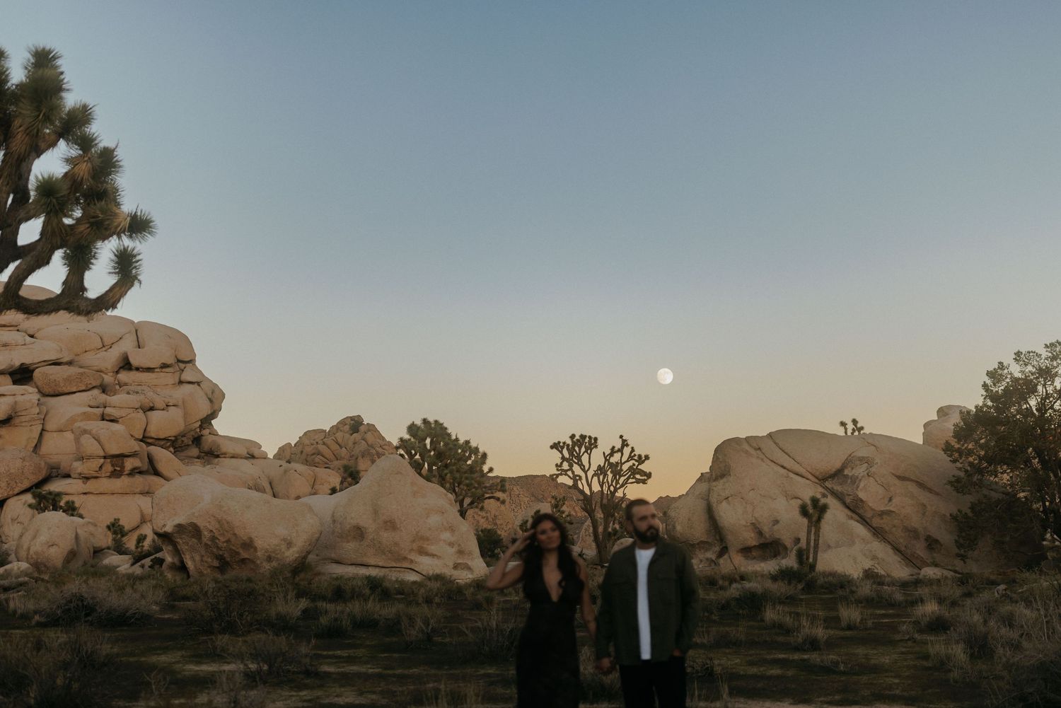 Couple standing together at dusk in Joshua Tree with large boulders and full moon in desert landscape.