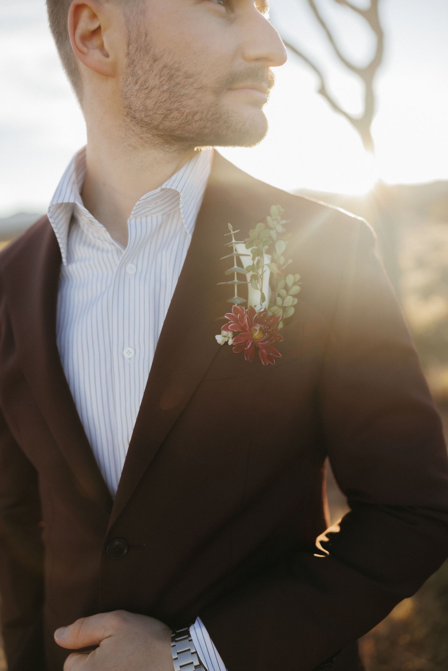 Groom in brown blazer with red floral boutonniere standing in warm golden sunset light.