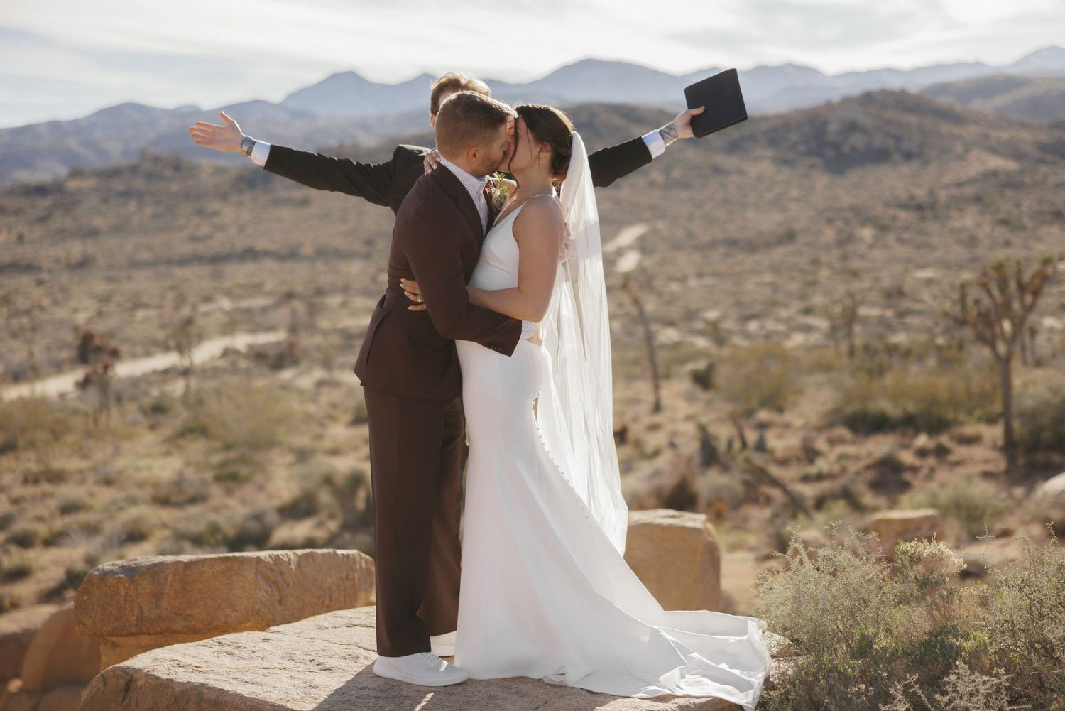 Newlyweds share a kiss on rocky overlook with sweeping desert mountain views behind them.