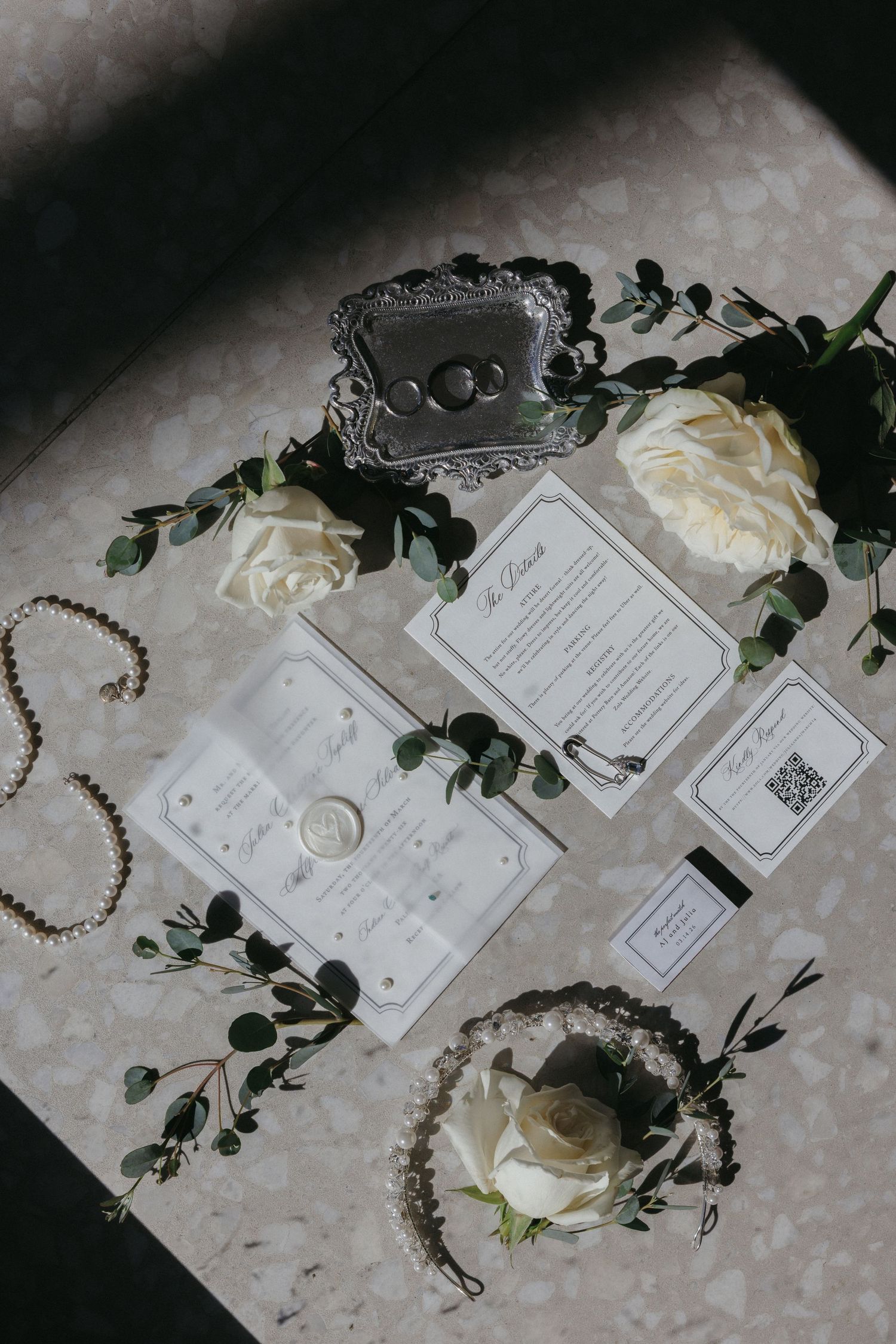 Elegant wedding flat lay with white roses, eucalyptus, rings, pearl jewelry, and stationery suite on marble surface.