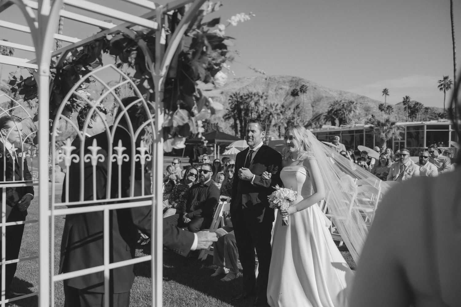 Bride walks down outdoor aisle with escort at elegant Palm Springs wedding ceremony.