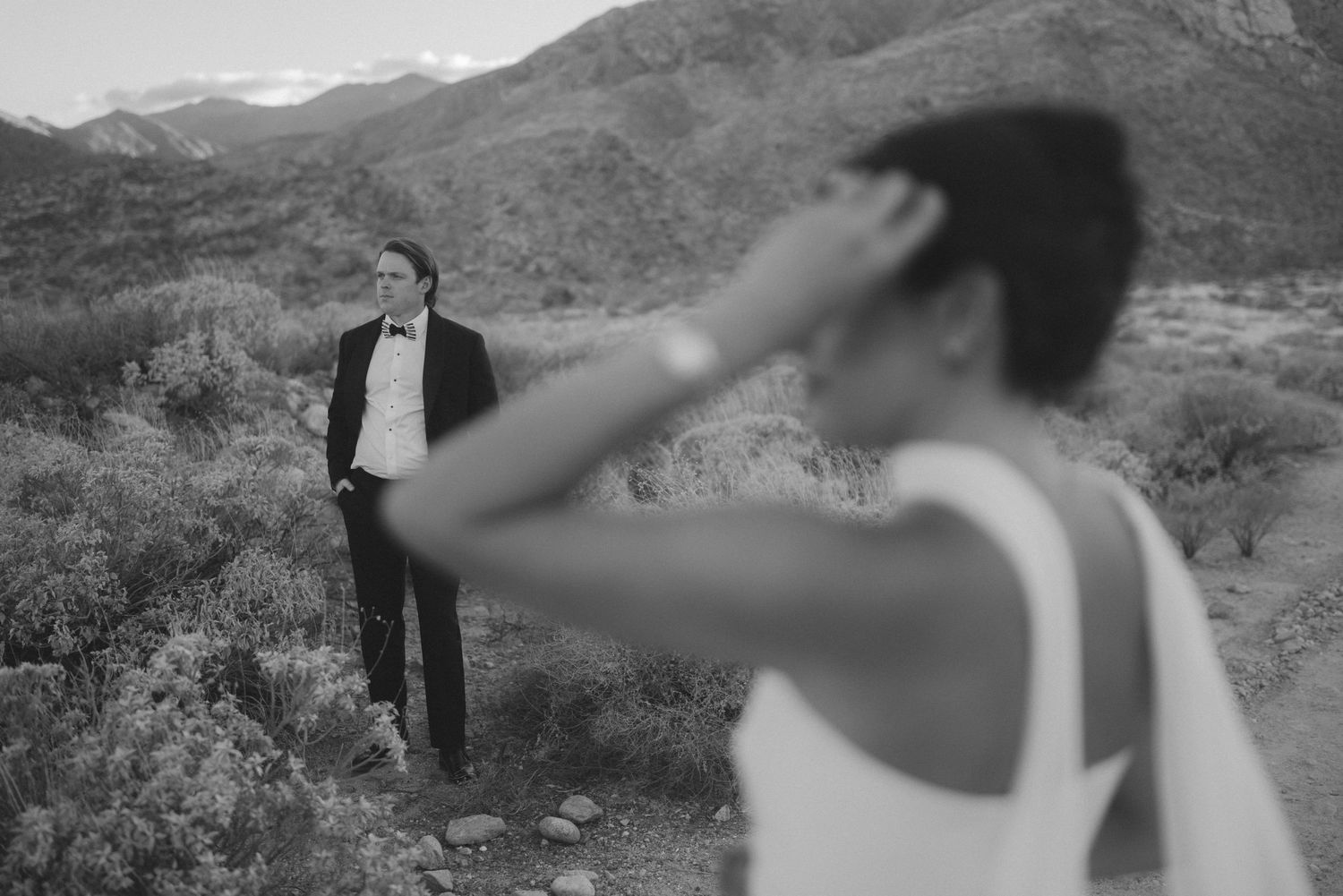 Groom in tuxedo stands in desert landscape as bride shields her eyes from the sun in black and white photo.