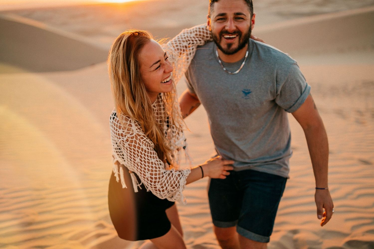 Couple laughing together on sand dunes during golden hour sunset in desert setting.