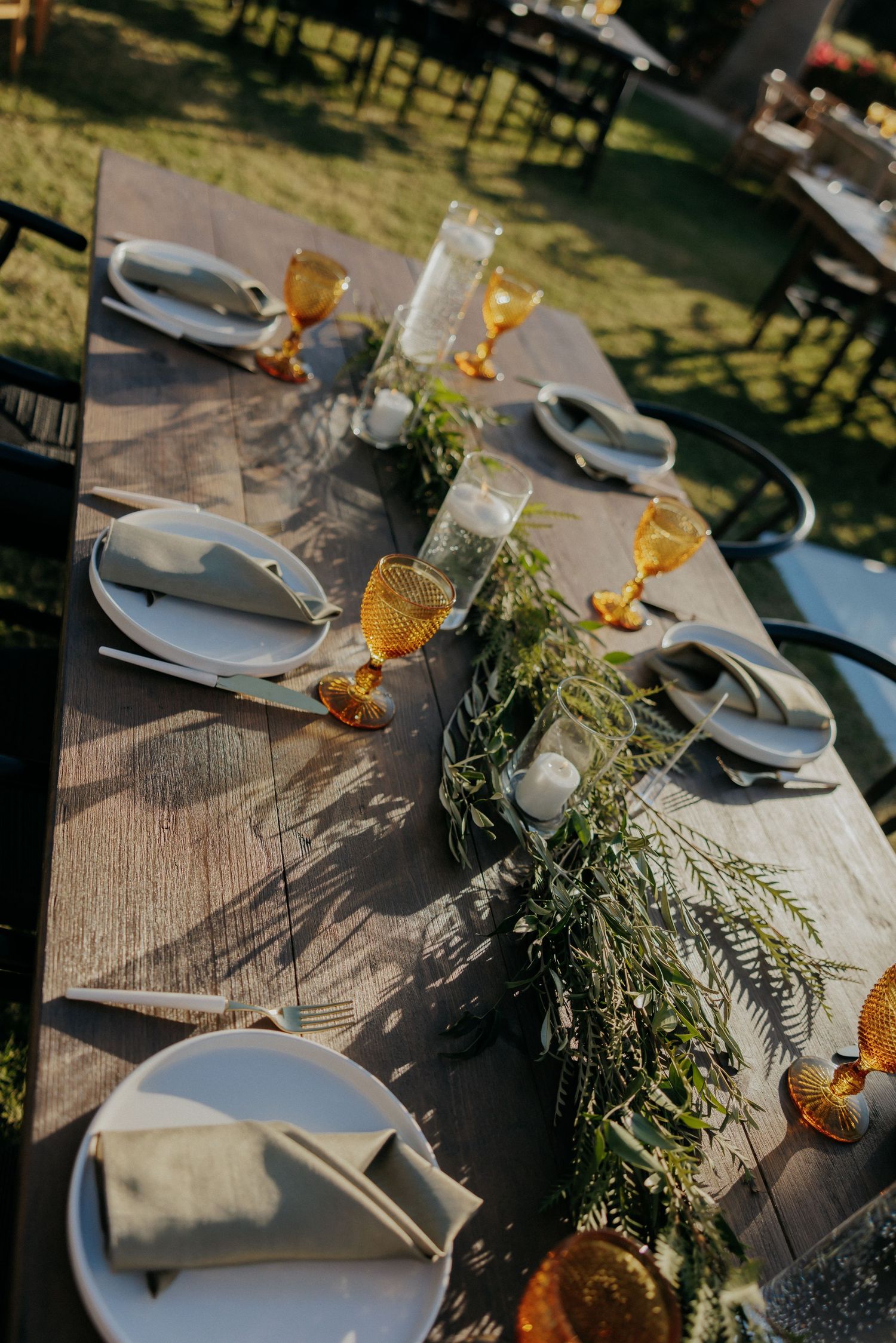 Rustic outdoor table setting with gray plates, linen napkins, and evergreen garland centerpiece captured in natural sunlight.