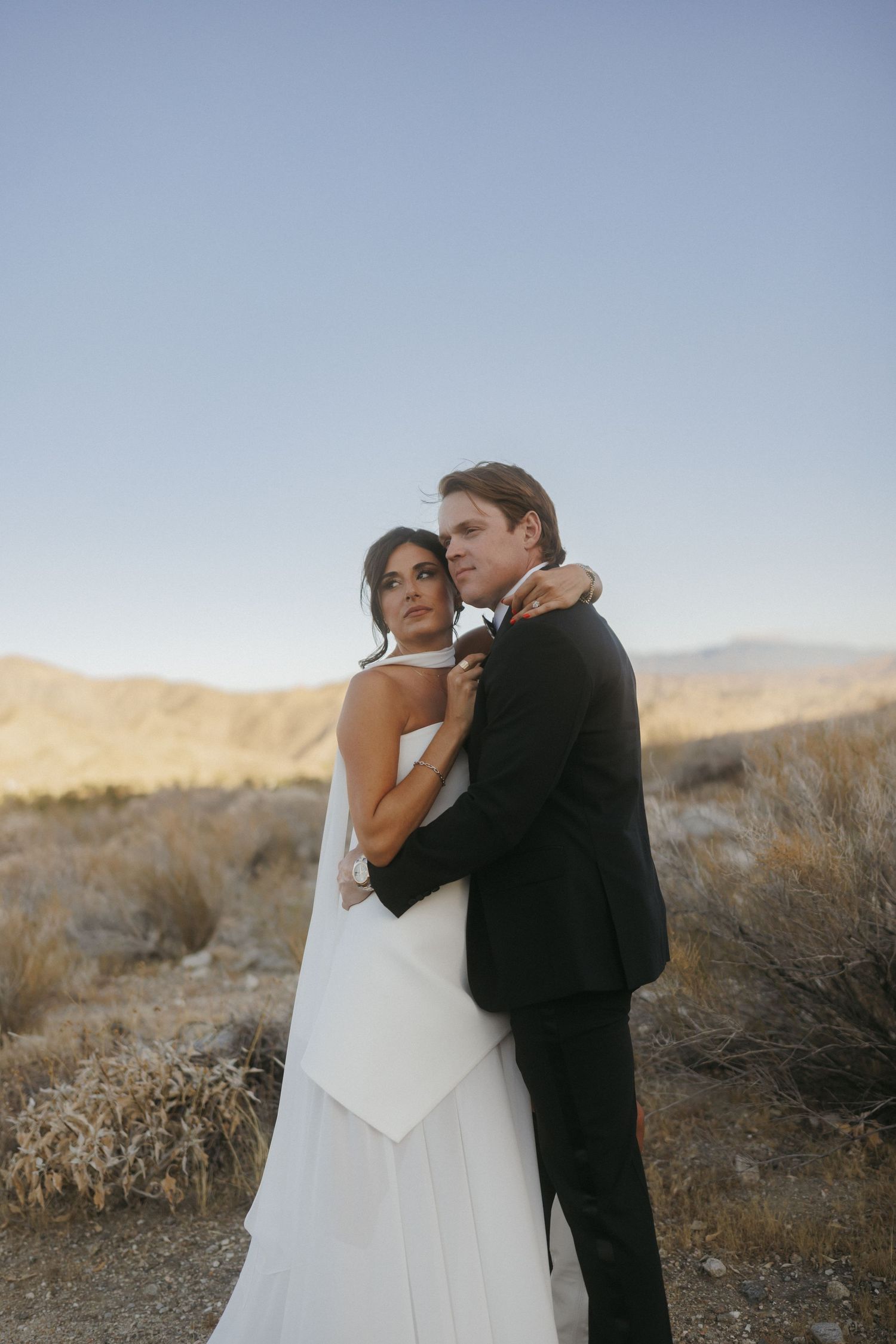 A bride and groom embrace in a desert landscape with mountains at sunset, she in a white gown, he in a black suit.