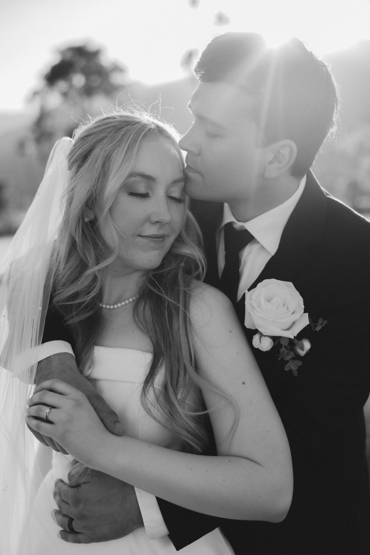 Black and white portrait of groom kissing bride on forehead in soft sunlight.
