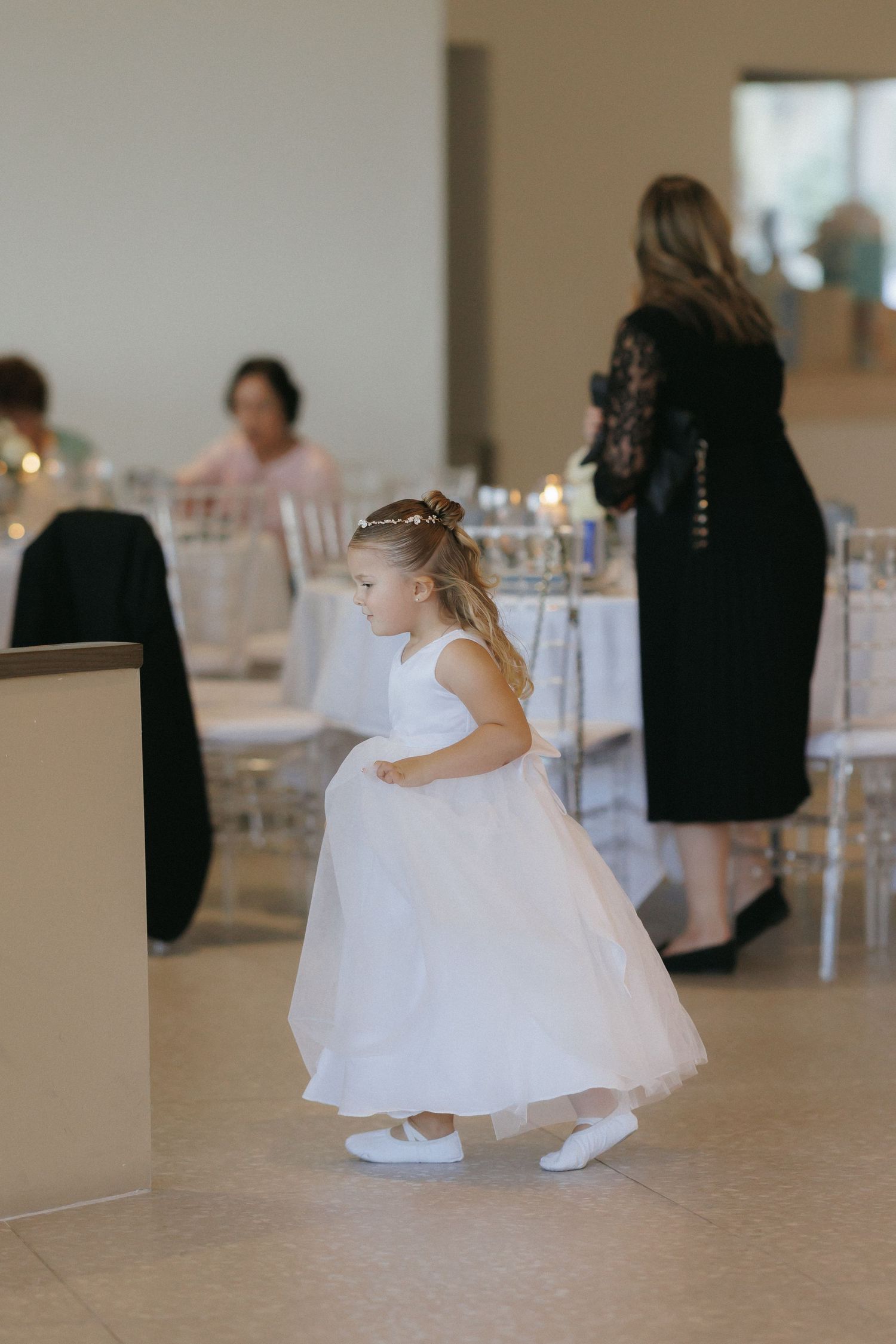 Young flower girl in white dress walking at elegant wedding reception with guests seated at candlelit tables.