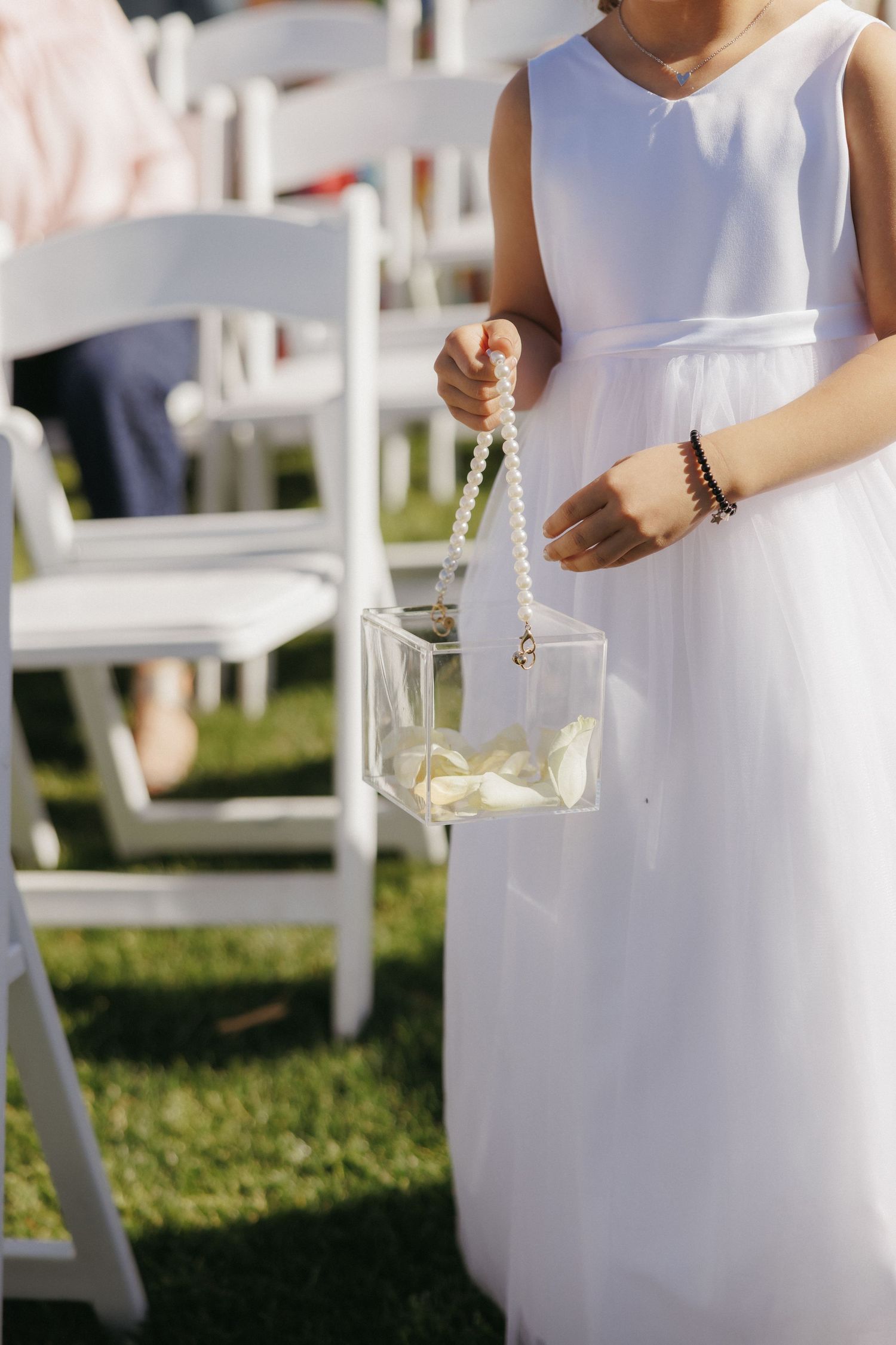 Flower girl in white dress holding a clear box with pearl handle filled with rose petals at outdoor wedding.