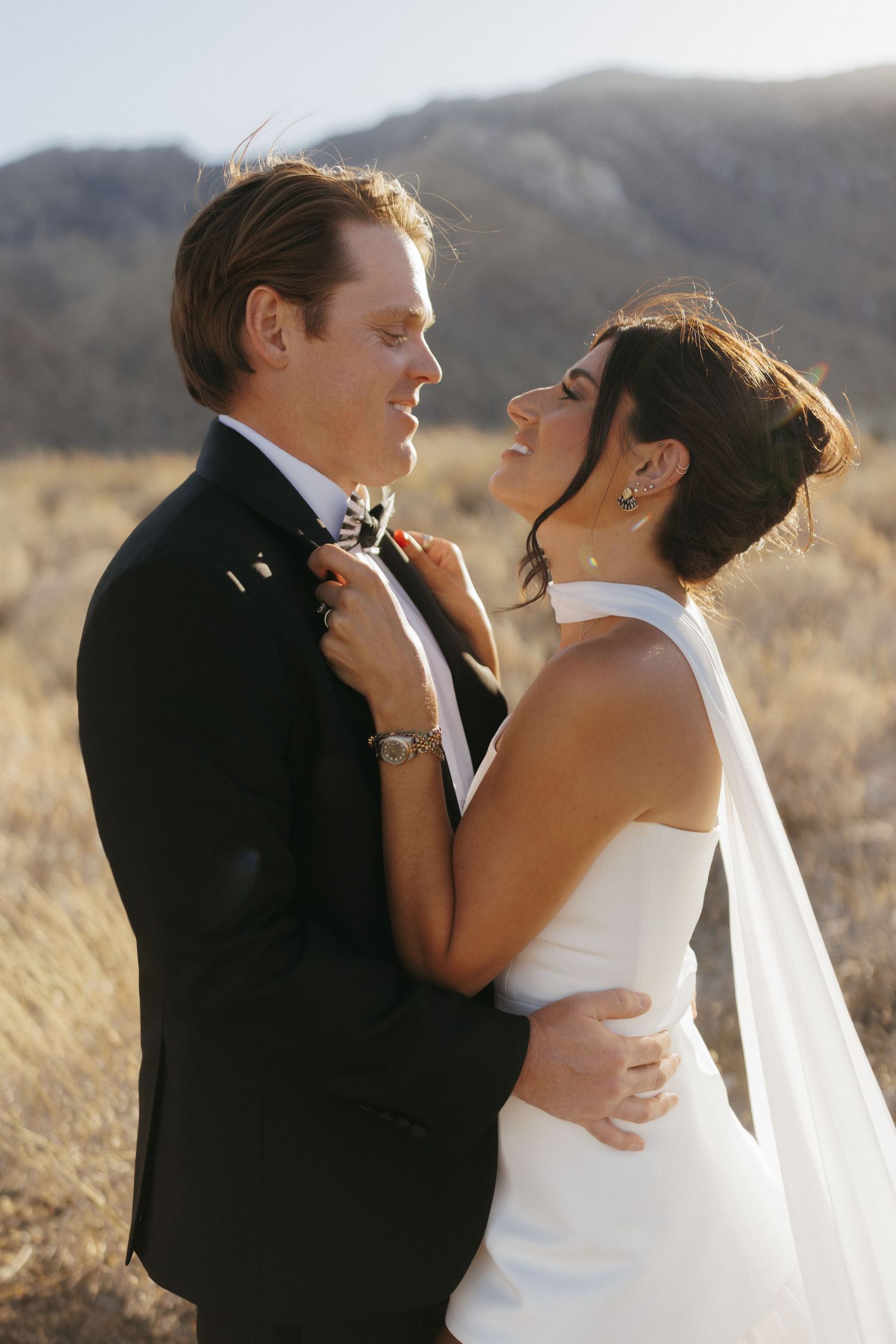 A bride and groom share a romantic moment outdoors with mountain scenery, she adjusts his bow tie while smiling.