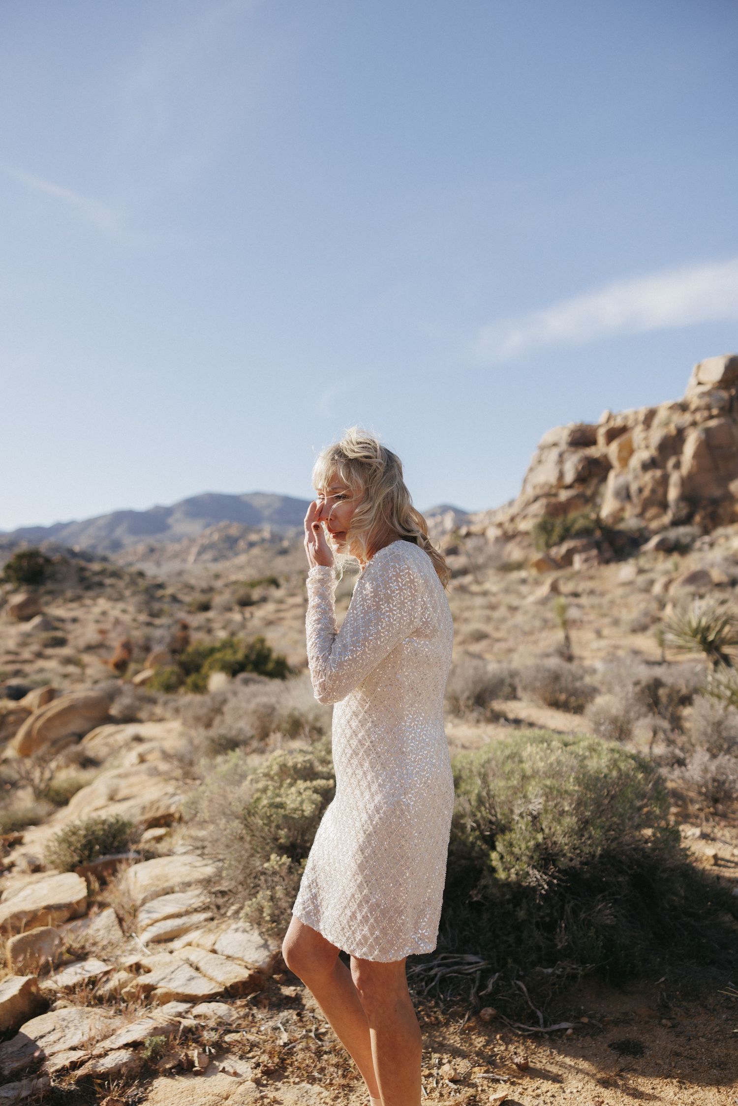 A blonde woman in a white lace dress stands in a rocky desert landscape under blue sky.