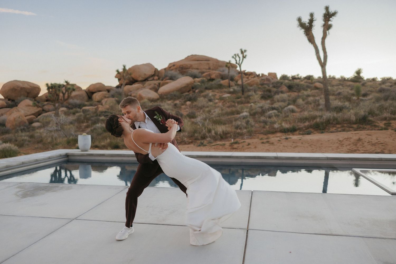 Groom dips bride in elegant white dress beside a pool with Joshua Tree boulder formations at dusk.