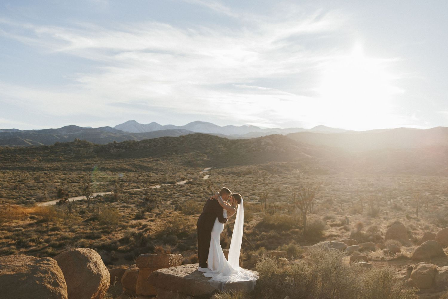 Bride and groom share a kiss on rocky overlook with sweeping desert mountain views.