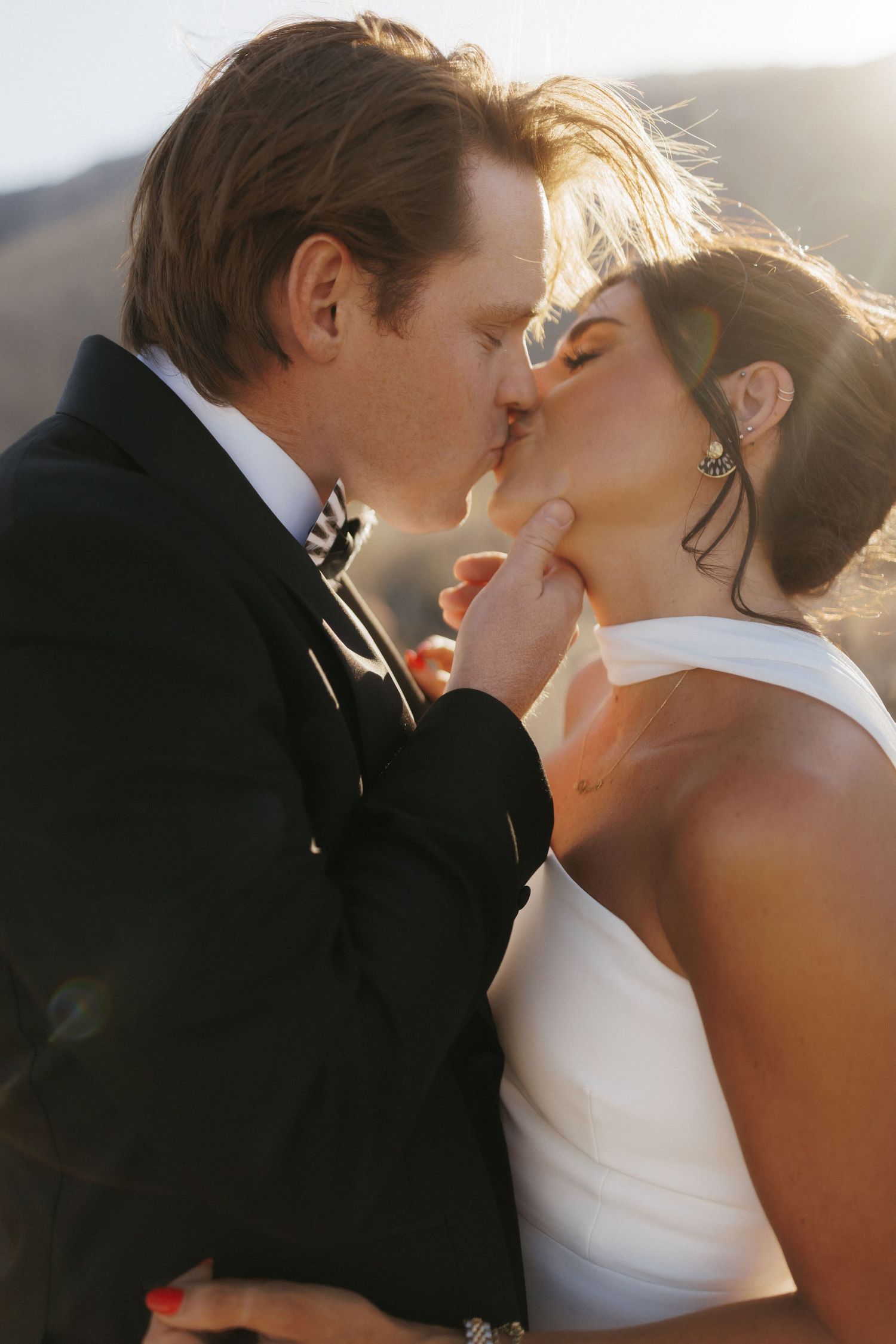 A bride and groom share a romantic kiss outdoors, with golden sunlight illuminating their wedding attire.