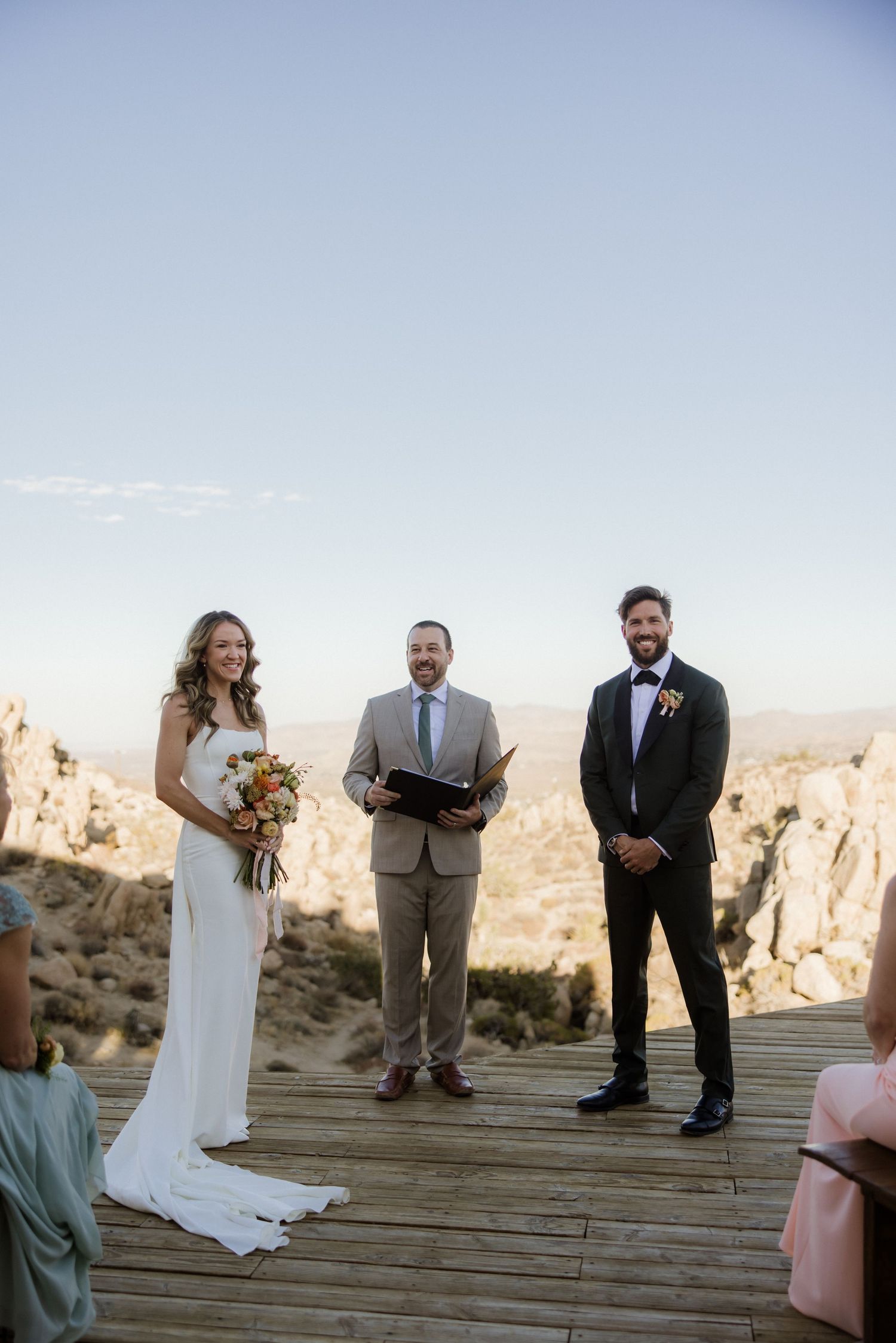 Wedding ceremony takes place on wooden deck in desert landscape with rocky mountains in background during sunset.