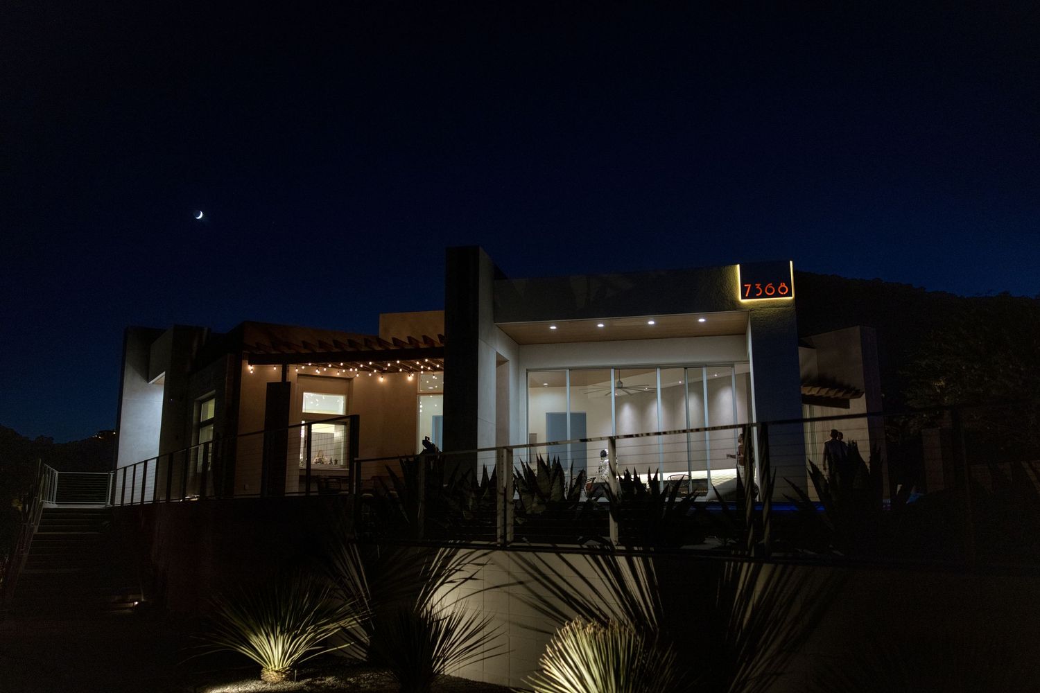 Modern house exterior illuminated with warm lighting against dark night sky showcases string lights and palm trees at dusk.