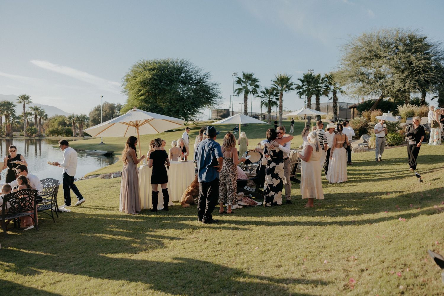 People gather on a grassy hillside lawn at an outdoor event with tents and palm trees in the background.