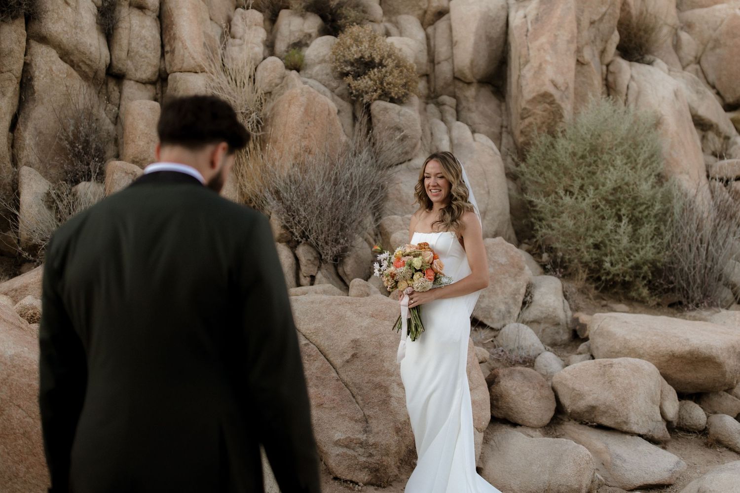 A couple in formal wedding attire pose together at Joshua Tree National Park among desert rock formations.