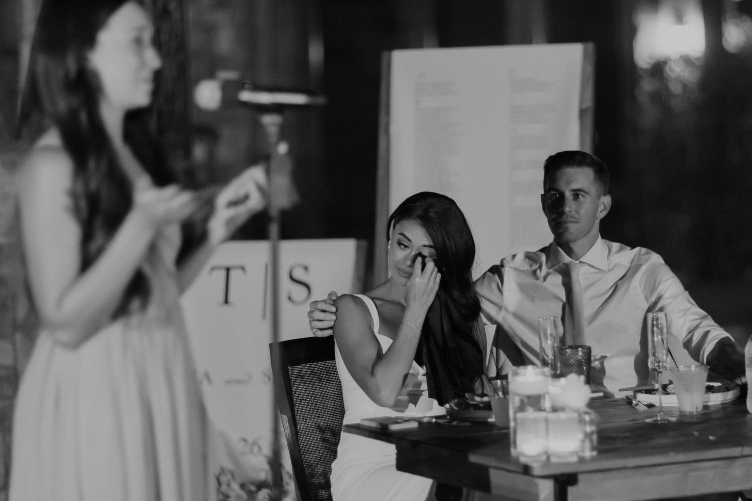 A candid black and white wedding reception scene with guests seated at tables while someone delivers a speech.