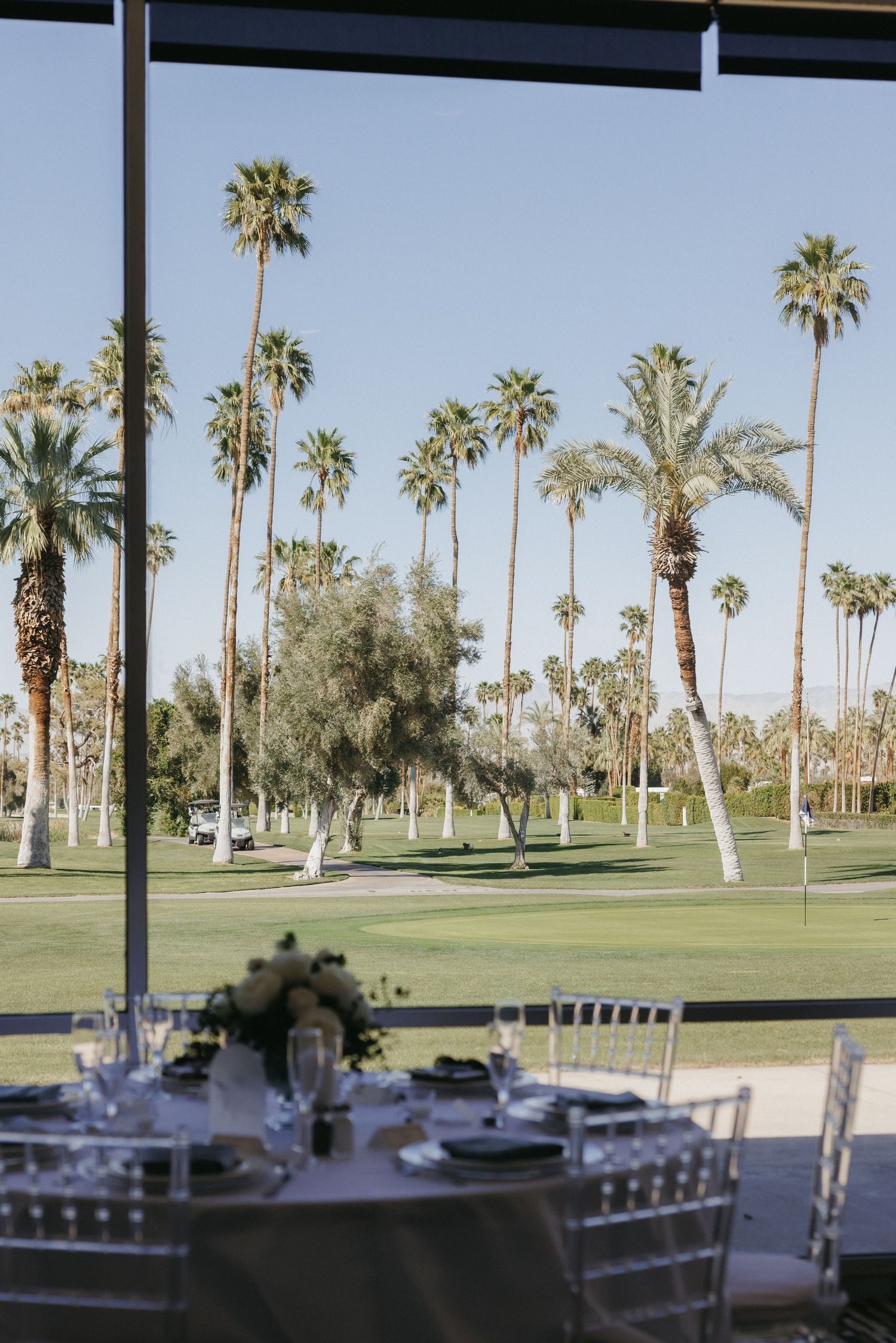 Elegant wedding reception table set near floor-to-ceiling windows overlooking a lush golf course lined with tall palm trees.