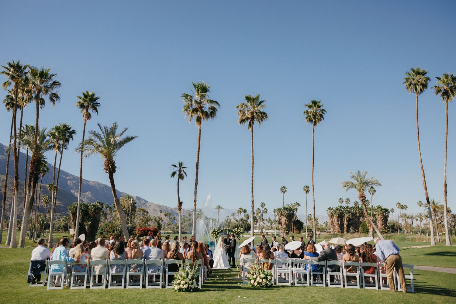Outdoor wedding ceremony on lush green lawn surrounded by tall palm trees with mountain backdrop in Palm Springs.