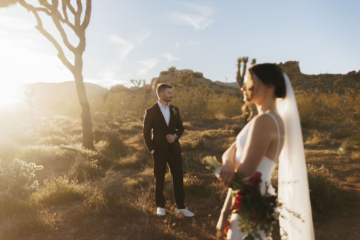 Groom in dark suit gazes at bride holding bouquet in sunlit desert.