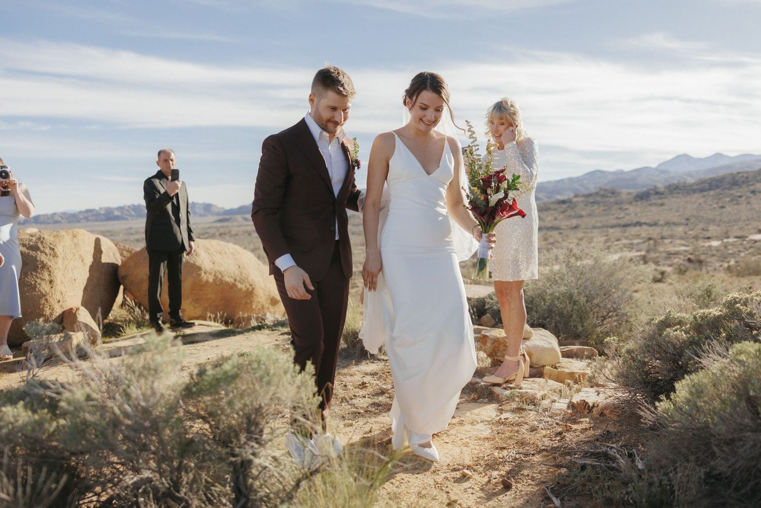 Bride and groom walk together through desert landscape during outdoor wedding ceremony with guests in background.