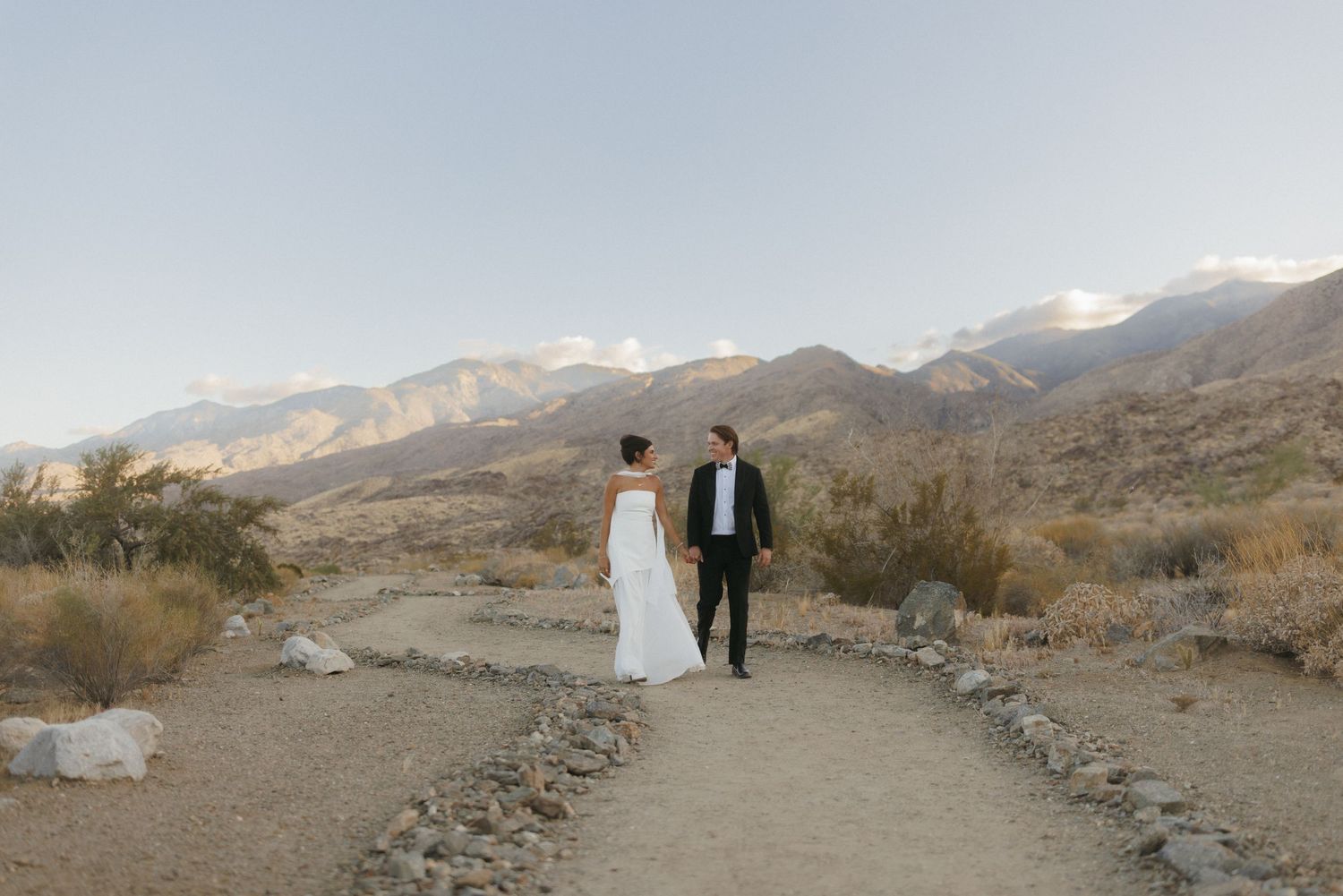 Bride and groom walking along a desert path with stunning mountain range backdrop at dusk.