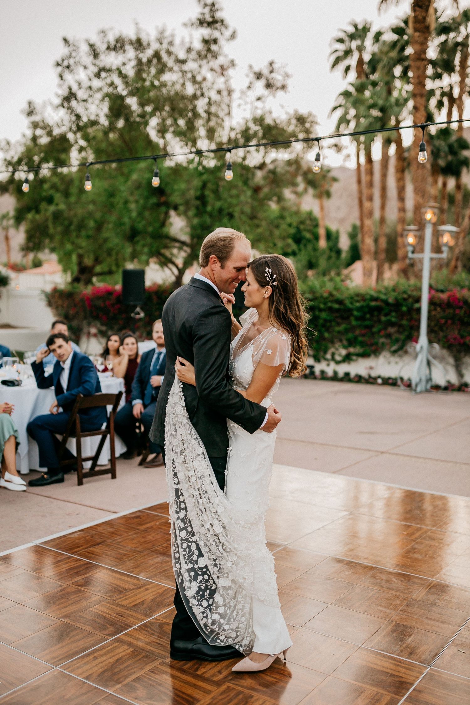 Couple dances on a wooden floor at an outdoor wedding reception with string lights overhead.