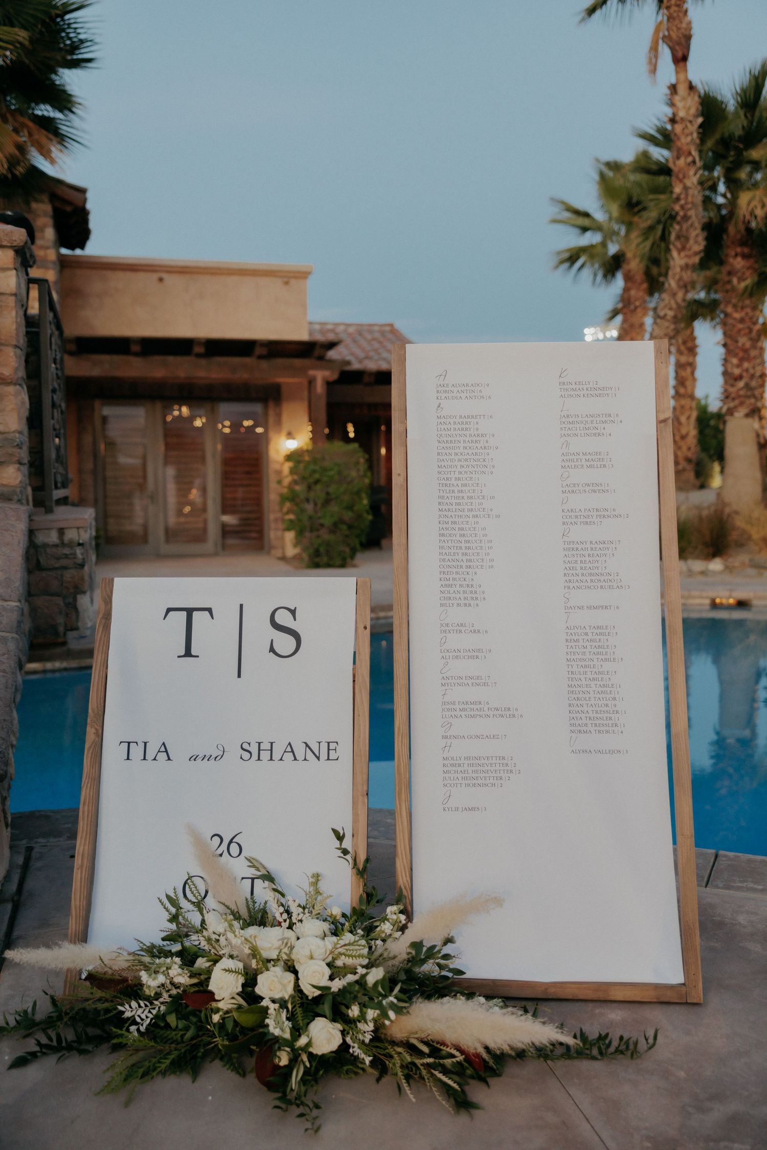 Elegant wedding seating chart display with floral arrangements and white paper signs against a poolside backdrop.