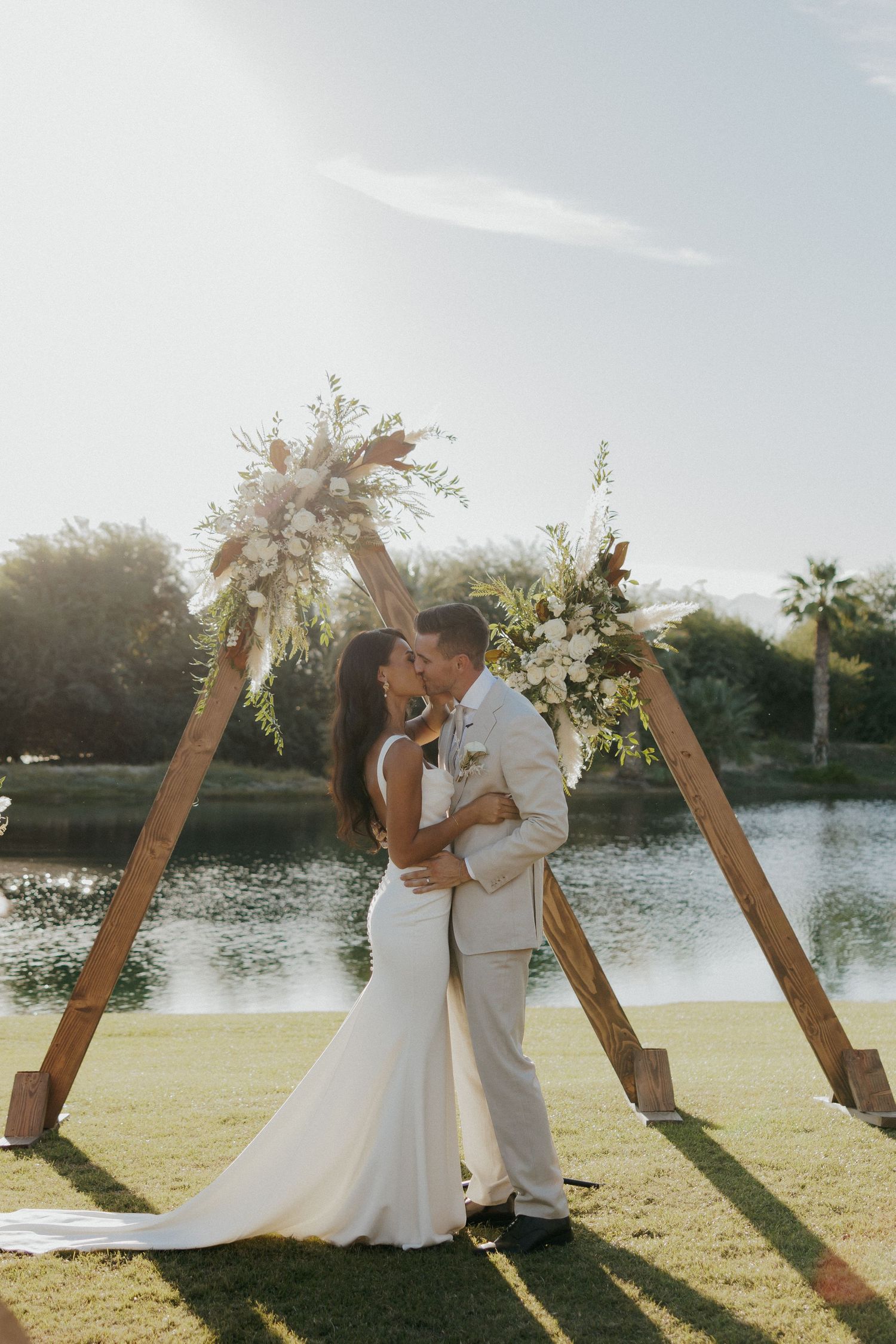 A couple embraces on their wedding day beside a serene lake with a wooden arch decorated in white flowers during sunset.