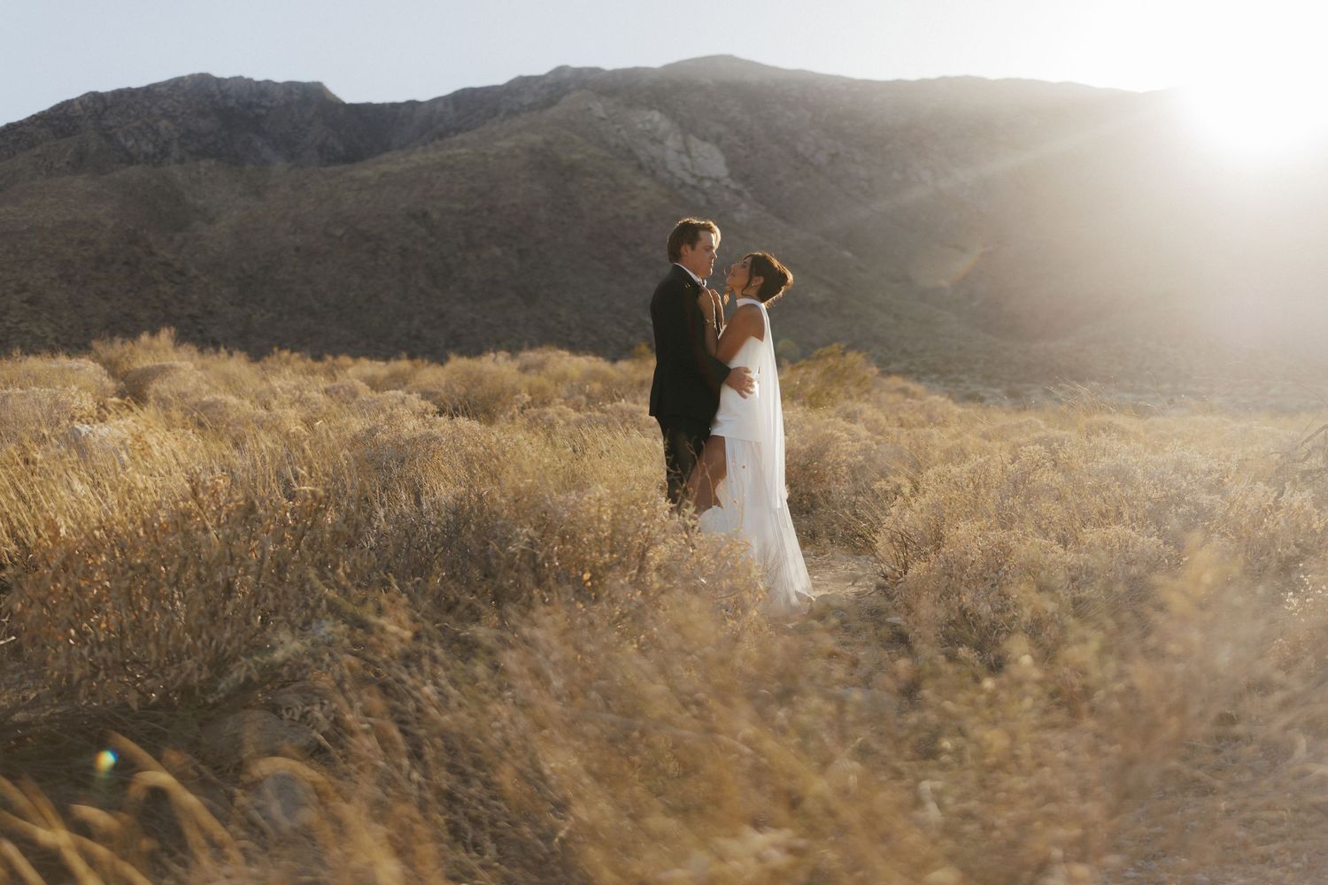 Bride and groom embrace in golden desert field with sunlit mountains backdrop during outdoor wedding ceremony.