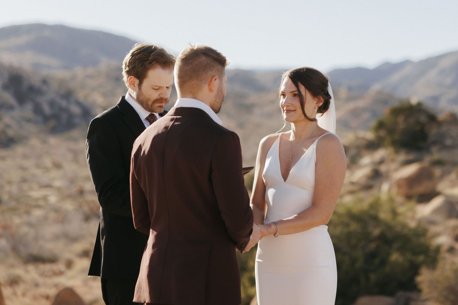 Couple exchanges wedding vows outdoors in desert landscape with officiant, mountains in background.